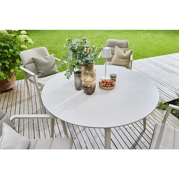 Sunfun Solo Gartentisch rund - Ø x H: 120 x 74 cm, Aluminium, Sintered Stone, Light-Greige - Ein runder Gartentisch mit Dekorationen wie Blumen und Schalen, umgeben von bequemen Stühlen auf einer Holzterrasse.