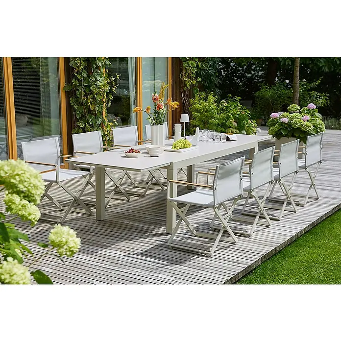 Sunfun Gartenmöbel-Set Maja/Solo - 9 -tlg., Light-Greige, Ausziehbar - Ein langer, weißer Gartentisch mit passenden Stühlen auf einer hölzernen Terrasse, umgeben von üppigem Grün. Perfekt für Mahlzeiten im Freien.