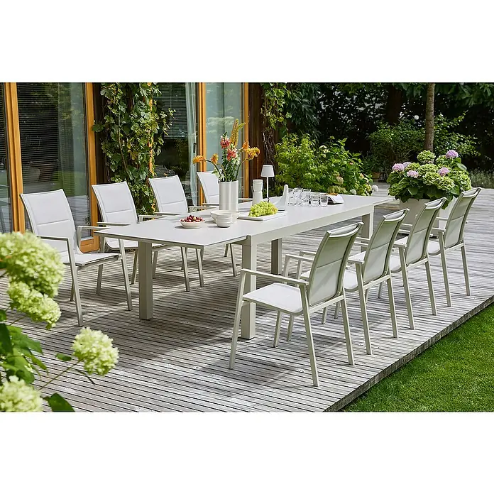 Sunfun Maja Gartenmöbel-Set Stone - 9 -tlg., Sintered Stone, Aluminium, Textilene, Light-Greige - Ein langer Esstisch mit passenden Stühlen im Freien. Das Tischset besteht aus einem Tisch, Stühlen, Blumen und Geschirr auf einer Holzterrasse.