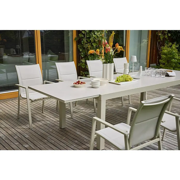 Sunfun Maja Gartentisch Stone - L x B x H: 300 x 100 x 74 cm, Aluminium, Sintered Stone, Ausziehbar, Light-Greige - Ein eleganter Esstisch mit weißen Stühlen auf einer Holzterrasse, dekoriert mit Obst, Blumen und stilvollen Accessoires für ein entspanntes Ambiente.