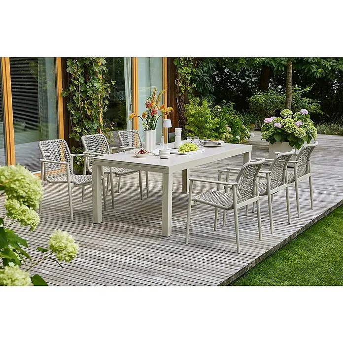 Sunfun Gartenmöbel-Set Maja/Solo - 7 -tlg., Light-Greige, Ausziehbar, Tisch (Lx B x H): 200-300 x 100 x 74 cm, Stuhl (B x T x H): 67 x 63 x 85 cm - Ein Esstisch-Set mit sechs Stühlen auf einer Holzterrasse, umgeben von üppigem Grün. Das Set lädt zum Verweilen im Freien ein.