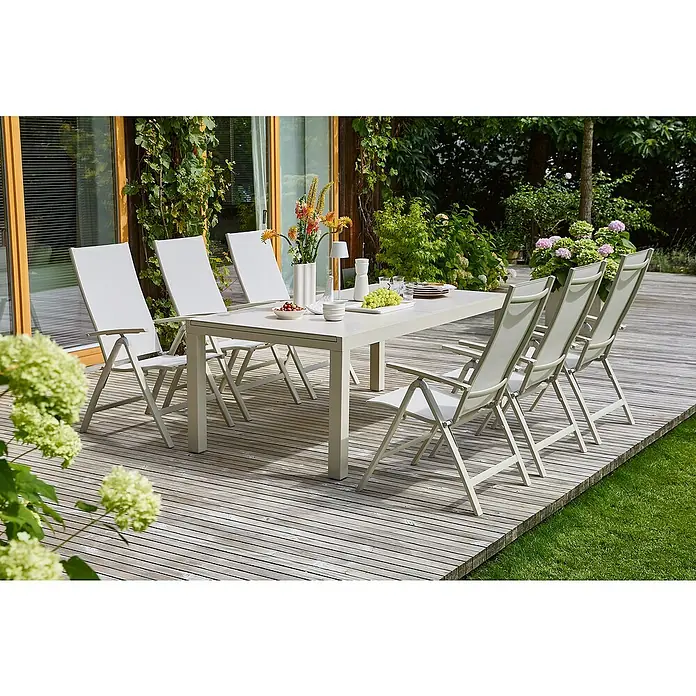 Sunfun Gartenmöbel-Set Maja - 7 -tlg., Aluminium, Sintered Stone, Textilene, Light-Greige - Ein Esstisch-Set im Freien mit einem hellgrauen Tisch und weißen Stühlen auf einer Holzterrasse, umgeben von üppigem Grün.