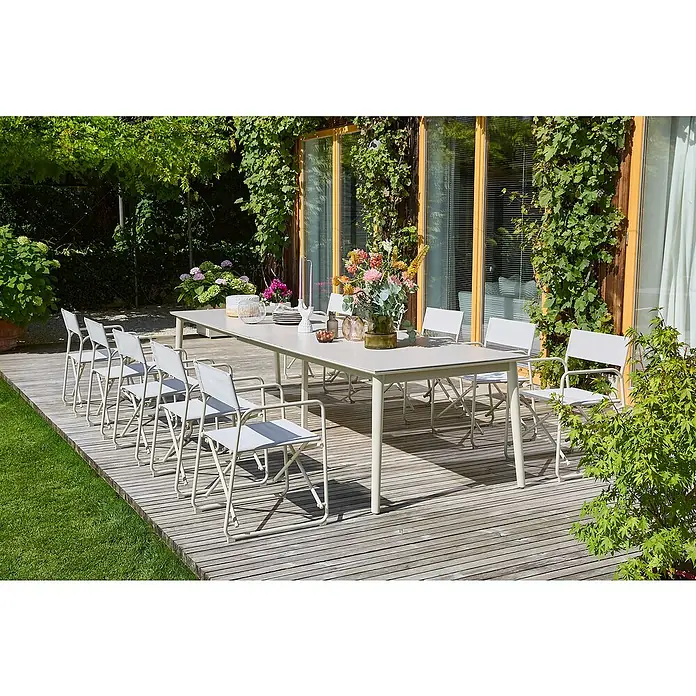 Sunfun Gartenmöbel-Set Solo/Sarah - 12 -tlg., Aluminium/Sintered Stone/Textilen, Light-Greige, Ausziehbar - Ein langer Esstisch mit passenden Stühlen steht auf einer Holzterrasse, umgeben von üppigem Grün. Perfekt für Mahlzeiten im Freien.