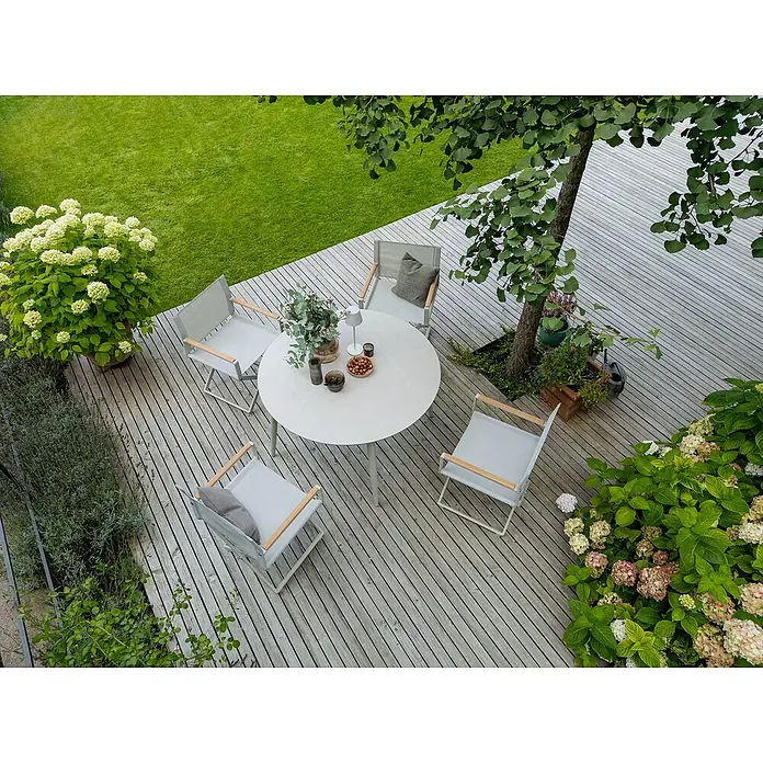 Sunfun Solo Gartentisch rund - Ø x H: 120 x 74 cm, Aluminium, Sintered Stone, Light-Greige - Gartensitzgruppe mit grauem Tisch und Stühlen auf einer Holzterrasse, umgeben von üppigem Grün und Pflanzen.