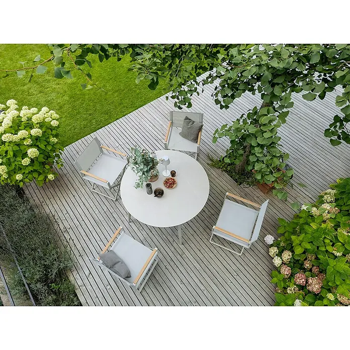 Sunfun Solo Gartentisch rund - Ø x H: 120 x 74 cm, Aluminium, Sintered Stone, Light-Greige - Ein runder Tisch mit vier Stühlen auf einer Terrasse, umgeben von üppigem Grün. Die Möbel laden zum Verweilen im Freien ein.