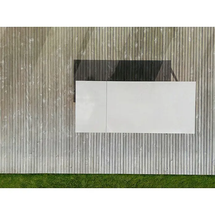 Sunfun Solo Gartentisch - L x B x H: 230 x 100 x 76,5 cm, Aluminium, Sintered Stone, Light-Greige, Ausziehbar - Eine rechteckige, weiße Tischplatte liegt auf einem Holzboden mit grüner Grasfläche. Der Tisch wirft einen dunklen Schatten.