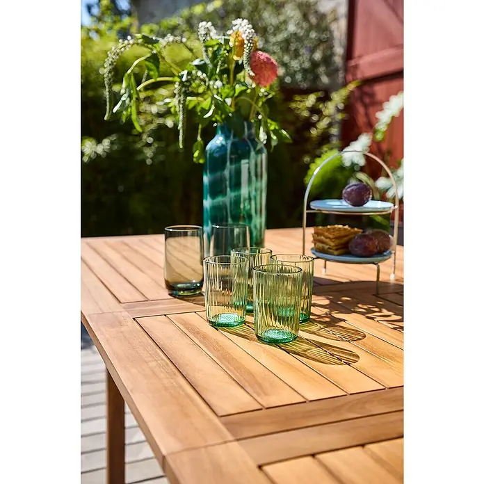 Sunfun Diana Gartentisch - L x B x H: 180 x 100 x 75 cm, Akazie, Naturbraun, Ausziehbar - Nahaufnahme von sechs Trinkgläsern auf einem Holztisch im Freien, umgeben von Blumen und Snacks.