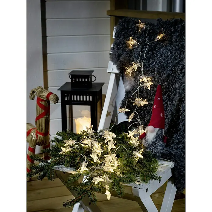 Konstsmide LED-Lichterkette Sterne - Außen, Lichtfarbe: Warmweiß, Anzahl Leuchtmittel: 40 Stk., Batteriebetrieben - Dekorative Sternenlichterkette auf einem weißen Stuhl mit Weihnachtsschmuck.