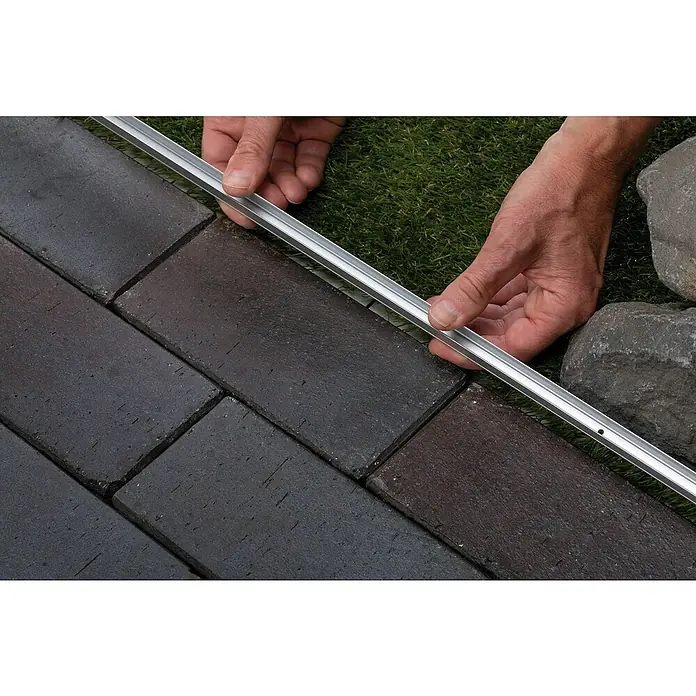 Paulmann Plug & Shine LED-Band Smooth Stripe Outdoor - Länge: 10 m, Lichtfarbe: Warmweiß, 1.540 lm, 24 V, 46 W - Hände installieren eine silberne Rasenkante neben dunklen Pflastersteinen und grünem Gras.
