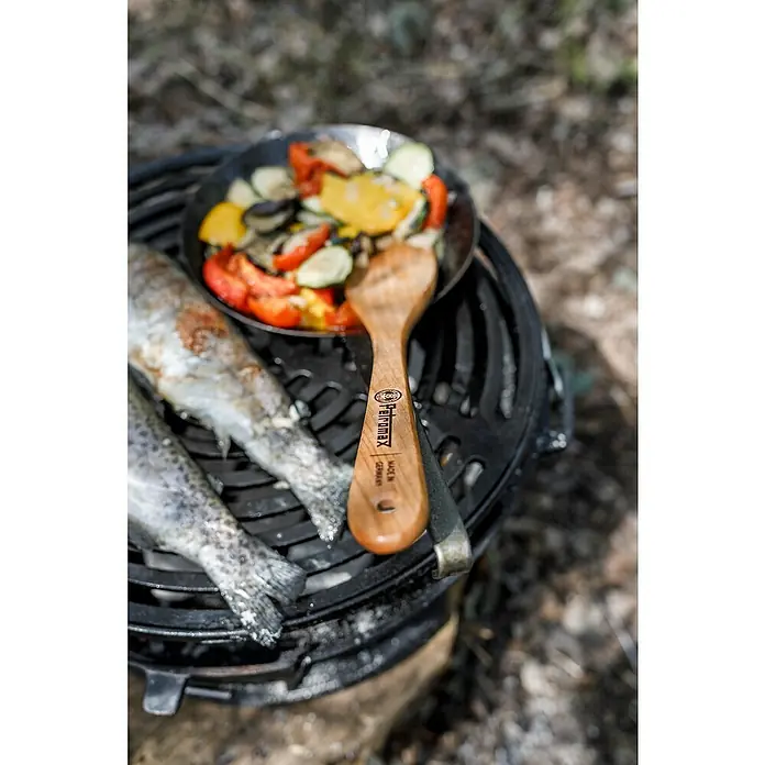Petromax Holzlöffel - Holz, Braun, 32 cm - Nahaufnahme eines Petromax Holzkochlöffels neben Fisch und Gemüse auf einem Grill, der ein Outdoor-Kocherlebnis verspricht.