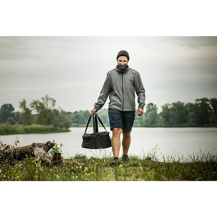 Petromax Transporttasche FT 12 - Ø x H: 45 x 24 cm, Polyester - Ein Mann trägt eine schwarze Tasche am Ufer eines Sees. Die vielseitige Tasche ist ideal für Outdoor-Abenteuer.