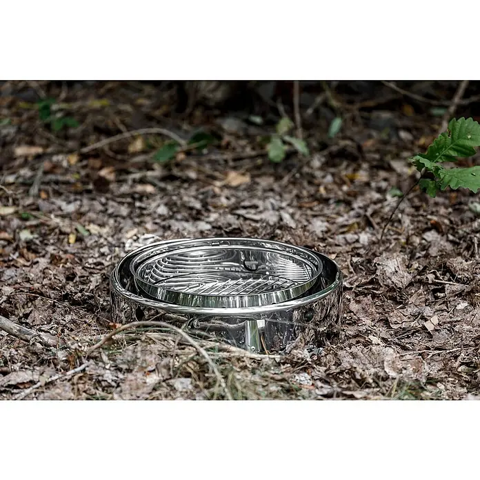 Petromax Holzkohlegrill Atago - Grillfläche (B x T): 45,1 x 44,5 cm, Durchmesser Grillfläche: 34 cm, Silber - Aufnahme eines glänzenden, tragbaren Outdoor-Kochers aus Edelstahl, der auf einer Laubstreufläche im Wald steht. Ideal für Camping und Outdoor-Abenteuer.