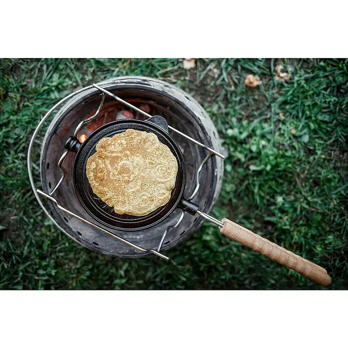 Petromax Waffeleisen zum Wenden - L x B: 54 x 22 cm, Gusseisen - Ein Pfannkuchen wird in einer gusseisernen Pfanne über einem Campingkocher zubereitet, umgeben von grüner Natur.