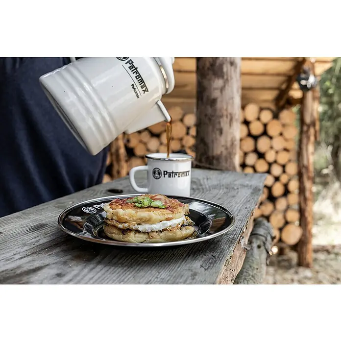 Petromax Teller rund - Ø x H: 22,5 x 2,5 cm, Schwarz, Stahl, 2 Stk. - Ein Petromax-Emaille-Kaffeekessel gießt Kaffee in eine Petromax-Tasse, dazu ein Frühstücksteller mit einem Sandwich auf einem Holztisch.