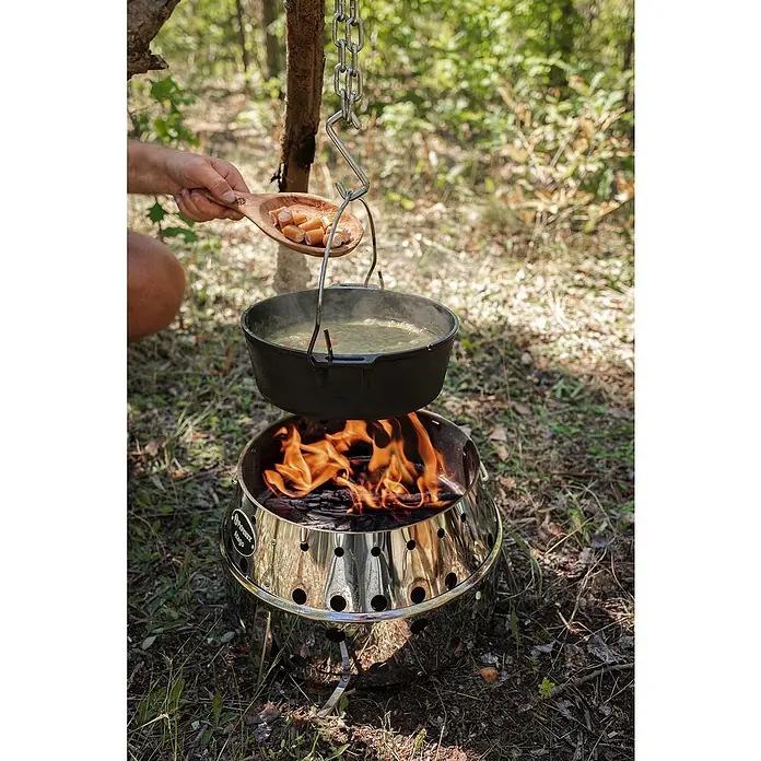 Petromax Holzkohlegrill Atago - Grillfläche (B x T): 45,1 x 44,5 cm, Durchmesser Grillfläche: 34 cm, Silber - Outdoor-Szene mit einem Dutch Oven über einem Lagerfeuer, jemand fügt Würstchen hinzu. Perfekt für Camping-Abenteuer!