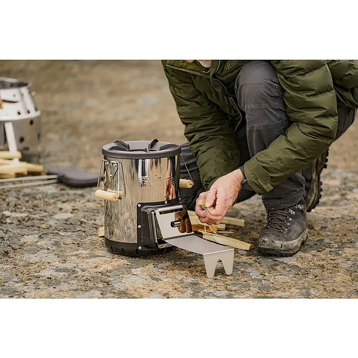 Petromax Holzkohlegrill Raketenofen - Grillfläche (B x T): 27,5 x 47,5 cm, Silber - Ein Mann befüllt einen tragbaren, silbernen Holzofen mit Holz. Ideal für Camping und Outdoor-Abenteuer.