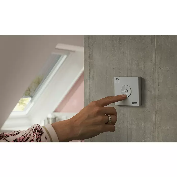Velux Funk-Wandschalter Hitzeschutz KLI 313 - Passend für: Außen liegende VELUX Solar-/Elektro-Rollläden und -Markisen (io-homecontrol®) - Eine Hand drückt eine weiße VELUX Wandbedienung mit Haussymbol und Richtungstasten, wahrscheinlich für Dachfenster.