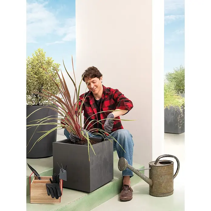 Deroma Millennium Macetero para plantas - Dimensiones externas (L x An x Al): 39 x 39 x 39 cm, Plástico, Antracita - Un hombre planta una planta en una maceta cuadrada de color antracita en una terraza, rodeado de herramientas de jardinería y vegetación.