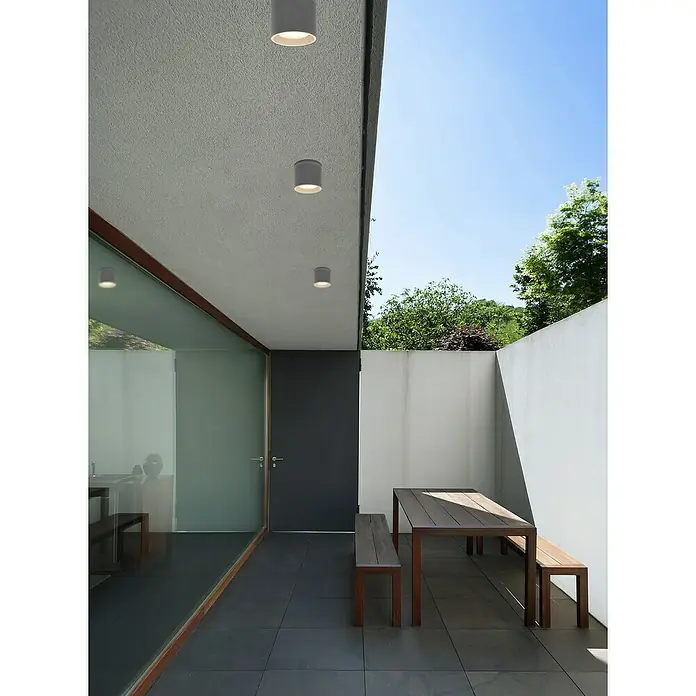 Globo Außenwandleuchte ALCALA - 11 W, Anthrazit, IP44 - Außenaufnahme eines modernen Gebäudes mit einer Terrasse, auf der eine Holzgarnitur und Deckenleuchten zu sehen sind.