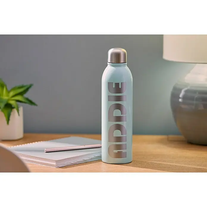 Cricut Vinylfolie Material Starter Bundle - 9 -tlg., Bunt - Hellblaue Wasserflasche mit silbernem Deckel und dem Aufdruck "Uddie", auf einem Holztisch, neben einem Notizbuch und einer Lampe.