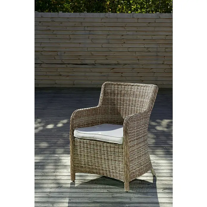 Sillón de jardín Gotland - An x Pr x Al: 79 x 63 x 99 cm, Arena - Primer plano de una silla de mimbre con un cojín claro sobre una plataforma de madera frente a una valla de madera.