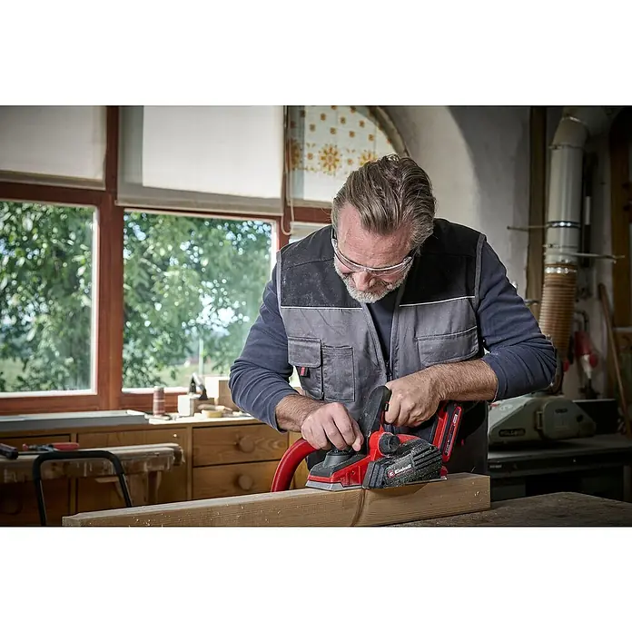 Einhell Power X-Change 18V Akku-Hobel TP-PL 18/3 LI BL - Solo - 18 V, Ohne Akku, Hobeltiefe: 3 mm, 12.000 U/min - Ein Mann benutzt einen Einhell Elektrohobel, um ein Stück Holz zu bearbeiten. Der Hobel ist rot und schwarz, und der Mann trägt eine Schutzbrille.