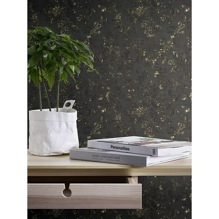 A.S. Création Neue Bude 2.0 ED II Vliestapete Damask - Schwarz/Gold, Ornament, 10,05 x 0,53 m - Nahaufnahme eines stilvollen Tisches mit einer Zimmerpflanze, Büchern und einer Schublade vor einer dunklen, floral gemusterten Tapete.