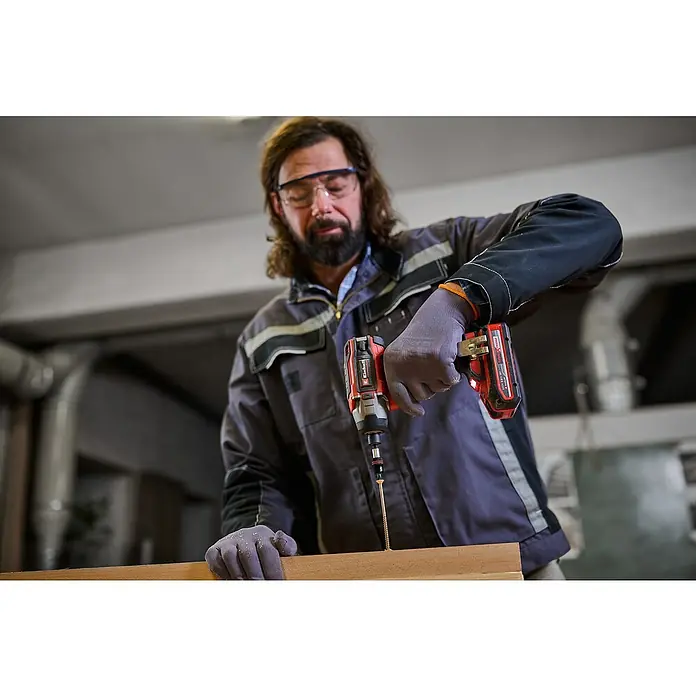 Einhell Professional Power X-Change 18V Akku-Schlagschrauber TP-CI 18/220 LI BL - 18 V, Ohne Akku, 220 Nm - Ein Handwerker mit Schutzbrille schraubt eine Schraube mit einem roten Akku-Bohrschrauber in ein Holzbrett.