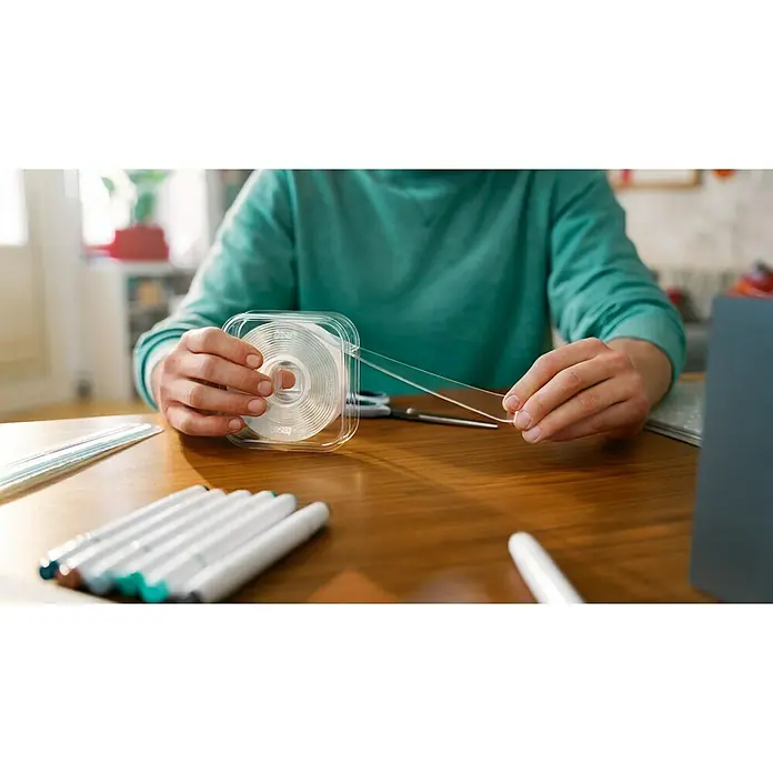 Tesa Doppelseitiges Klebeband Universal Tape with Nano Power - 3 m x 30 mm - Hände ziehen ein Stück transparentes, wiederverwendbares Klebeband aus der Spule. Der Klebestreifen ist in einer durchsichtigen Kunststoffbox.