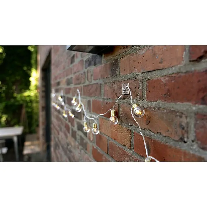 Tesa Doppelseitiges Klebeband Universal Tape with Nano Power - 3 m x 30 mm - Die Lichterkette mit den kleinen Glühbirnen hängt an einer roten Backsteinmauer und sorgt für eine gemütliche Atmosphäre im Außenbereich.