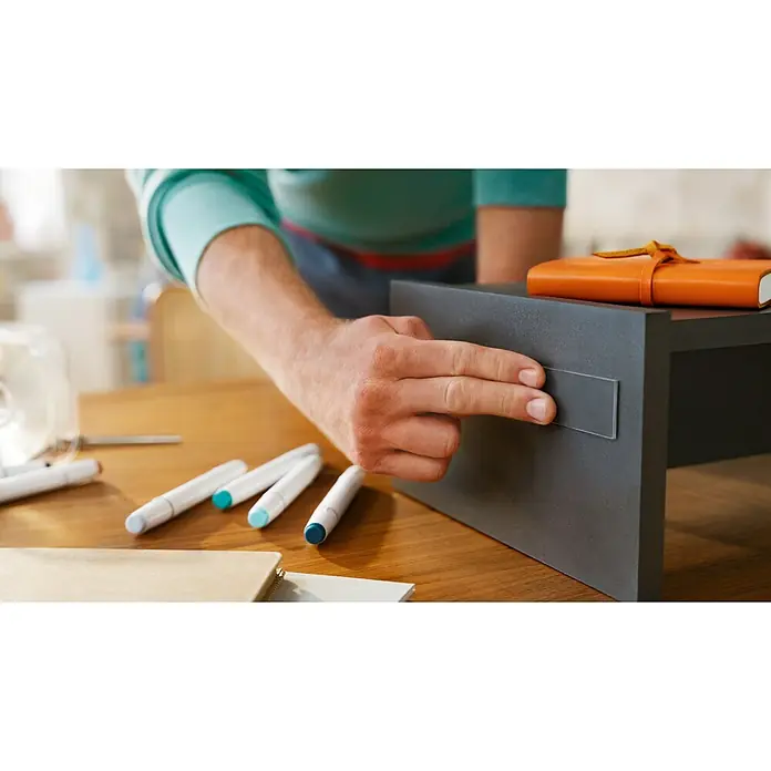 Tesa Doppelseitiges Klebeband Universal Tape with Nano Power - 3 m x 30 mm - Ein Mann befestigt einen Magnetstreifen an einem kleinen, dunklen Möbelstück auf einem Holztisch, um Gegenstände daran zu befestigen.