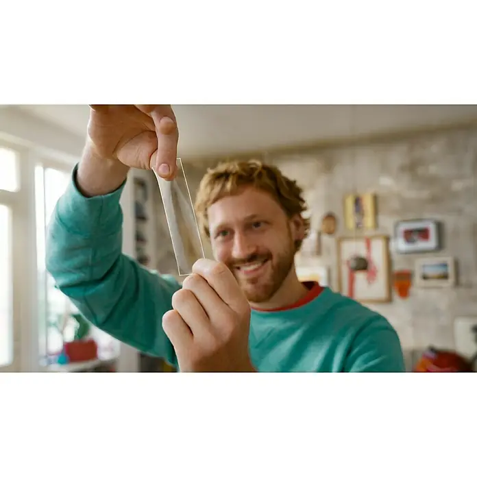 Tesa Doppelseitiges Klebeband Universal Tape with Nano Power - 3 m x 30 mm - Ein Mann hält eine dünne, transparente Glasplatte hoch, die das Licht reflektiert, während er lächelt. Im Hintergrund sind Kunstwerke zu sehen.