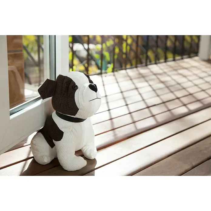 Rei Tope de puerta Perrito - Textil - Un peluche de perro con pelaje marrón y blanco está sentado en un suelo de madera soleado junto a una puerta corredera de cristal. Accesorio bonito y decorativo.