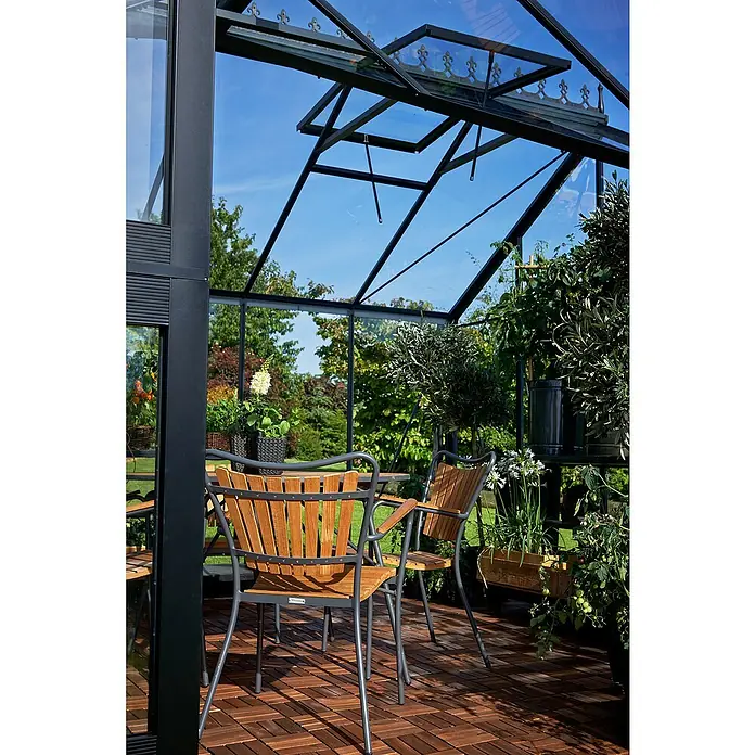 Juliana Gewächshaus Orangerie - Außenmaß inkl. Dachüberstand (B x T): 439 x 373 cm, Material Seitenwand: Glas, 3 mm, Anthrazit/Schwarz - Gemütliche Sitzecke im Gewächshaus mit Holzstühlen und Pflanzen, die eine entspannende Atmosphäre für ruhige Momente schaffen.