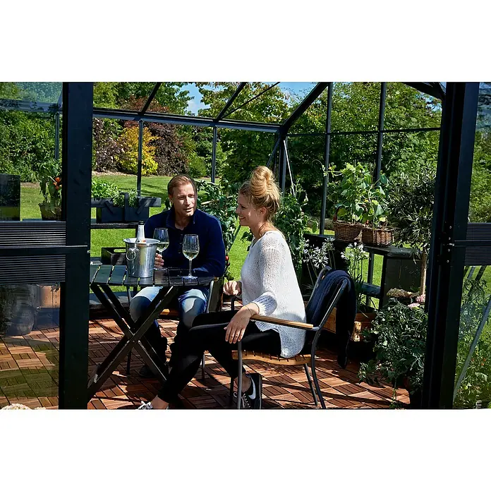 Juliana Gewächshaus Orangerie - Außenmaß inkl. Dachüberstand (B x T): 439 x 373 cm, Material Seitenwand: Glas, 3 mm, Anthrazit/Schwarz - Ein Gewächshaus mit einem Paar beim Weintrinken an einem Tisch inmitten üppiger Pflanzen. Entspannung und Genuss im eigenen Garten.
