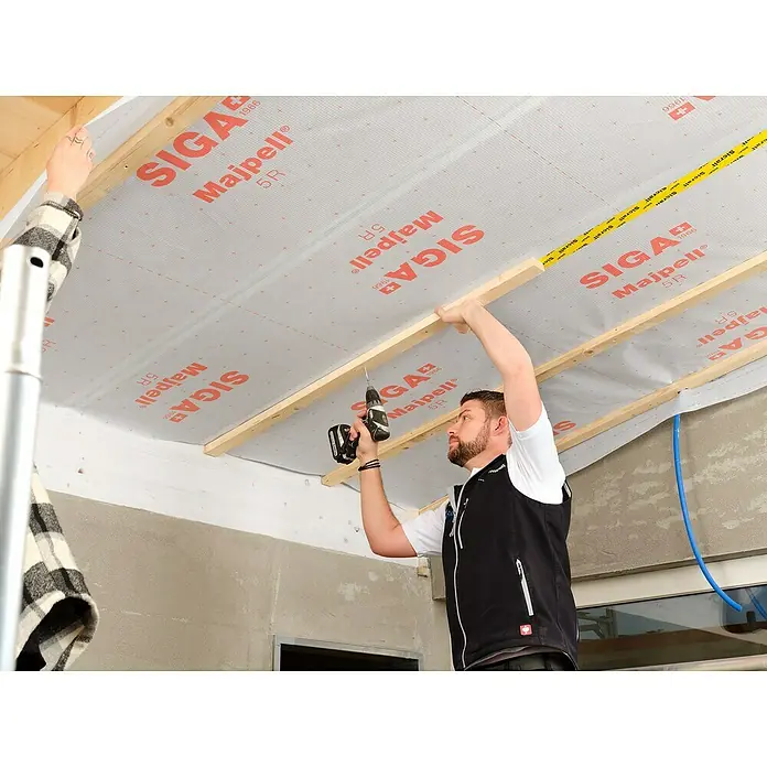 Siga Dampfbremsfolie Majpell 5 R - 30 m², Polyolefin - Ein Handwerker befestigt SIGA Majpell 5R-Dampfbremsfolie an einer Holzkonstruktion mit einem Akkuschrauber. Die Folie ist mit roter Schrift bedruckt.