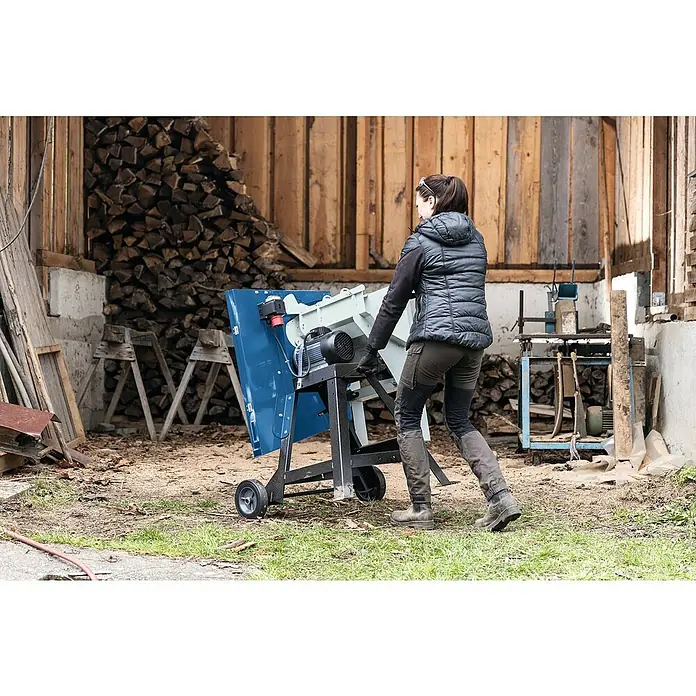 Scheppach Wippkreissäge HS720 - 4.500 W, Durchmesser Sägeblatt: 700 mm - Frau bewegt Kreissäge im Freien. Die mobile Kreissäge ist blau, schwarz und weiß und steht auf einem unbefestigten Untergrund.