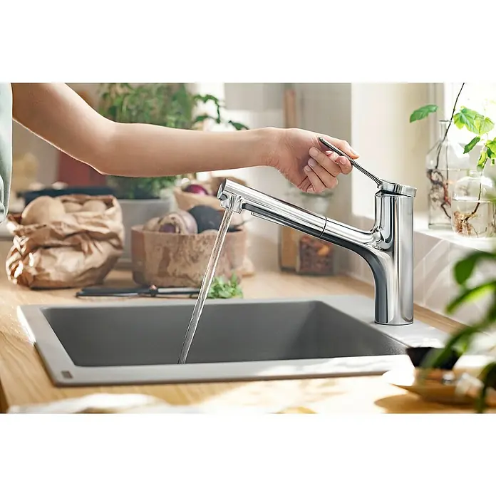 Hansgrohe Zesis M33 Spültischarmatur - Chrom, Glänzend, Mit sBox - Eine Hand betätigt einen modernen Chrom-Wasserhahn, aus dem Wasser in ein graues Spülbecken fließt. Im Hintergrund sind Küchenutensilien sichtbar.