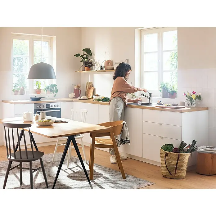 Hansgrohe Zesis M33 Spültischarmatur - Chrom, Glänzend, Mit sBox - Helle Küche mit Esstisch, Stühlen und einer Frau am Waschbecken, die ein Glas Wasser füllt. Eine einladende und funktionale Raumatmosphäre.