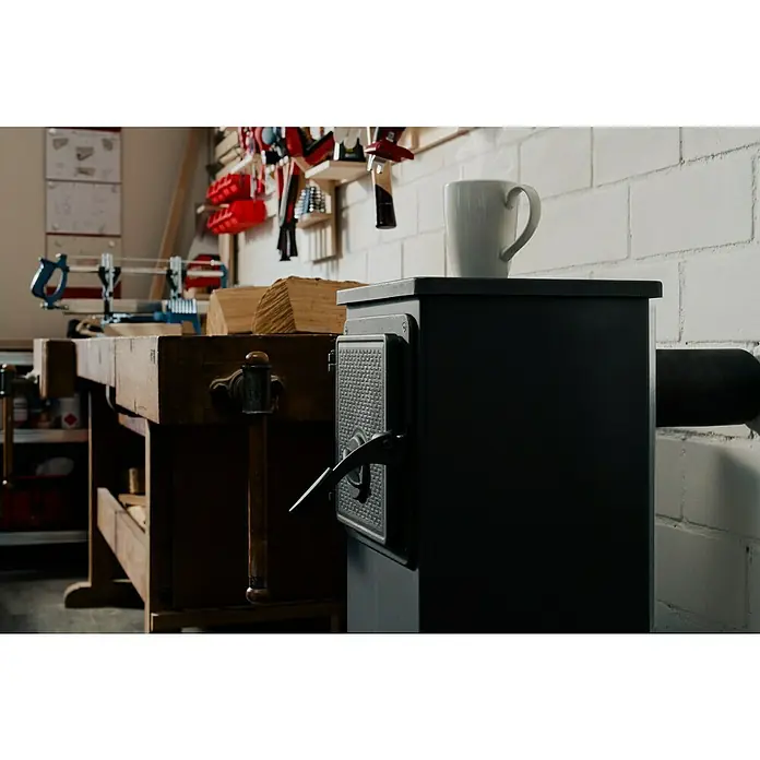 Werkstattofen Thalia - 5,3 kW, Raumheizvermögen: 160 m³, Schwarz - Nahaufnahme eines schwarzen Ofens mit einer weißen Tasse darauf, daneben eine Werkbank mit Holz, Werkzeugen an der Wand.