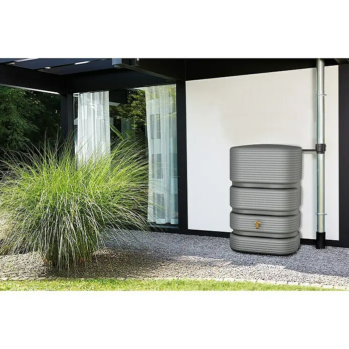 4rain Regenspeicher Quadro - 500 l, Betongrau - Grauer, horizontal gerippter Regenspeicher mit Messinghahn an einer weißen Wand neben einem Fallrohr.