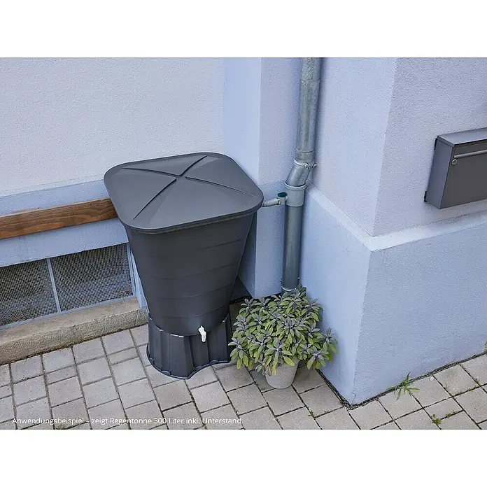 Garantia Regentonne Ibiza - 300 l, Graphitgrau, 1 Stk. - Regentonne im Garten. Die graue Tonne steht auf Steinplatten neben einem Fallrohr und einem Fenster.