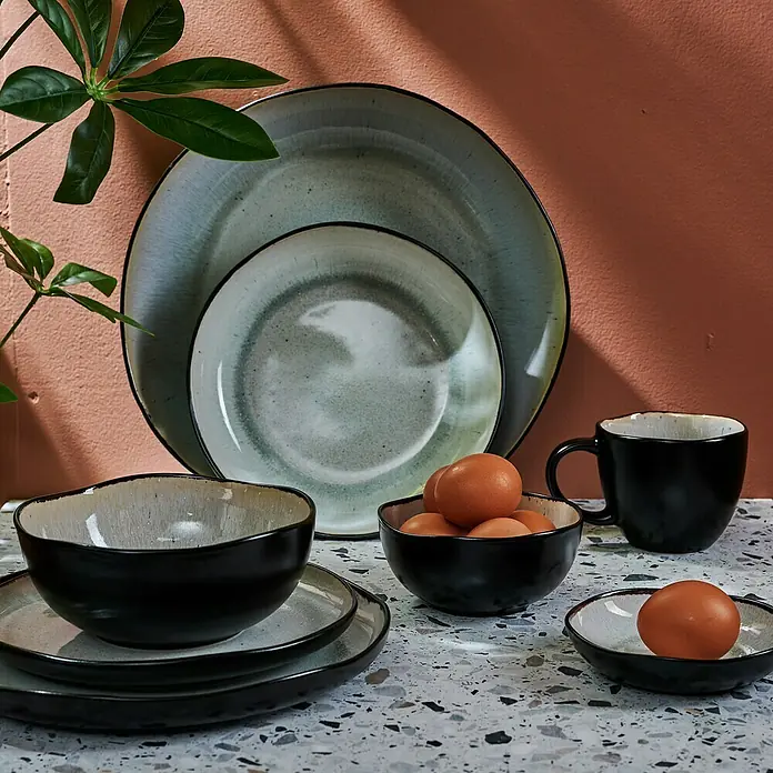 Teller Tief Lunar - 1 Stk., 22,5 x 5 cm, Keramik - Nahaufnahme von handgefertigtem Geschirrset in Erdtönen, bestehend aus Tellern, Schüsseln und einer Tasse, arrangiert auf einer Terrazzo-Oberfläche.