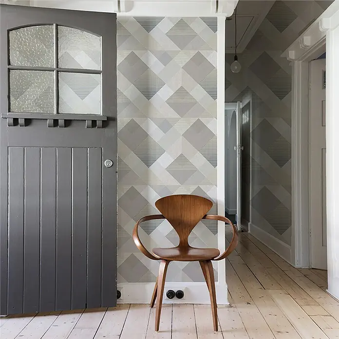 Papel pintado Geométrico rombos - Gris, 10 x 0,53 m - Un interior con una elegante silla de madera frente a una pared con un patrón geométrico, con una puerta gris adyacente.