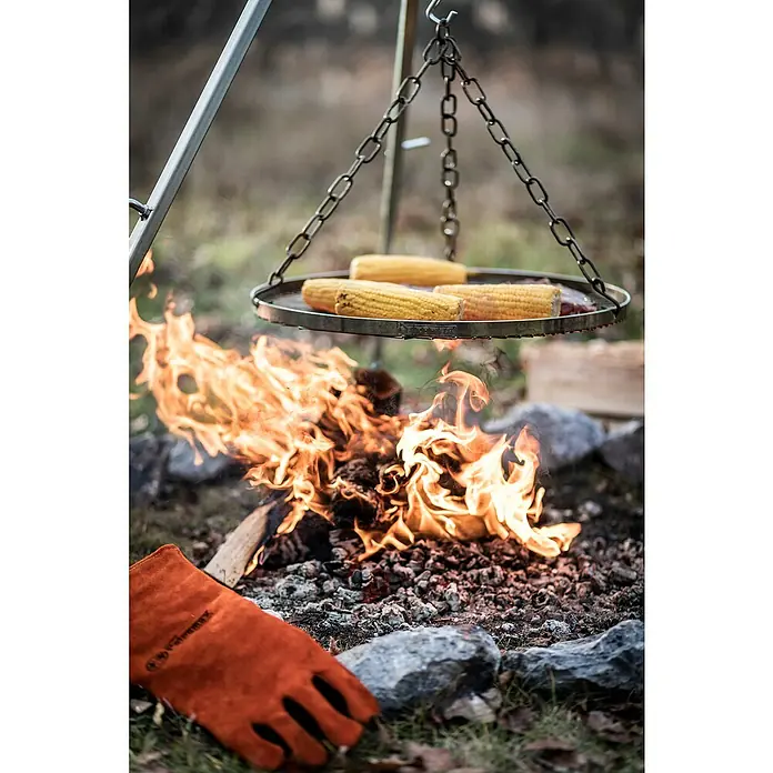 Petromax Grillrost Dreibeinständer - Durchmesser: 50 cm, Stahl - Ein Petromax Grillgalgen mit Maiskolben über einem Lagerfeuer, inklusive eines orangefarbenen Handschuhs. Perfekt für das Outdoor-Kochen und Grillen.