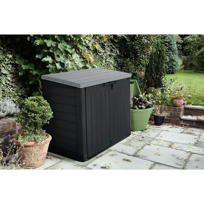 Keter Truhe Store it out Max - L x B x H: 145,5 x 82 x 125 cm, Anthrazit/Grau - Eine robuste, schwarze Gartenbox mit grauem Deckel steht auf einer gepflasterten Fläche, umgeben von üppigem Grün. Ideal zur Aufbewahrung von Gartengeräten.