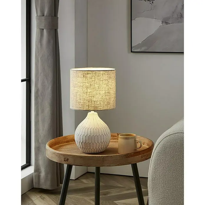 Lámpara de sobremesa redonda Bellariva 3 - 40 W, Ø x Al: 30 x 36 cm, Blanco, E27 - Primer plano de una lámpara de mesa decorativa con pantalla y base de cerámica sobre una mesa auxiliar, junto a una taza y cortinas.