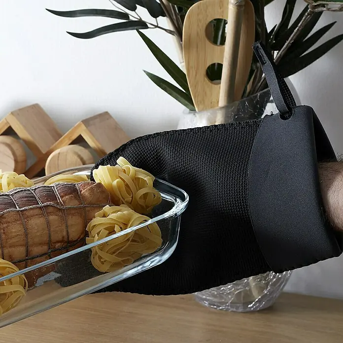 Manopla de neopreno - Goma, Negro/Burdeus, Negro/Gris, Negro/Beige - Un guante de horno negro sostiene una fuente de vidrio con asado y pasta, exhibiendo utensilios de cocina y plantas de interior como decoración.