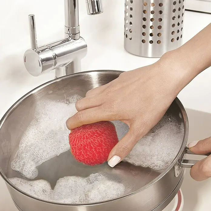 Estropajo antirrayadura - Plástico, 2 ud., Azul/Verde/Lila/Rosa - Una mano lava una olla llena de burbujas de jabón con una esponja roja, mientras que un grifo y un escurridor de utensilios son visibles al fondo.