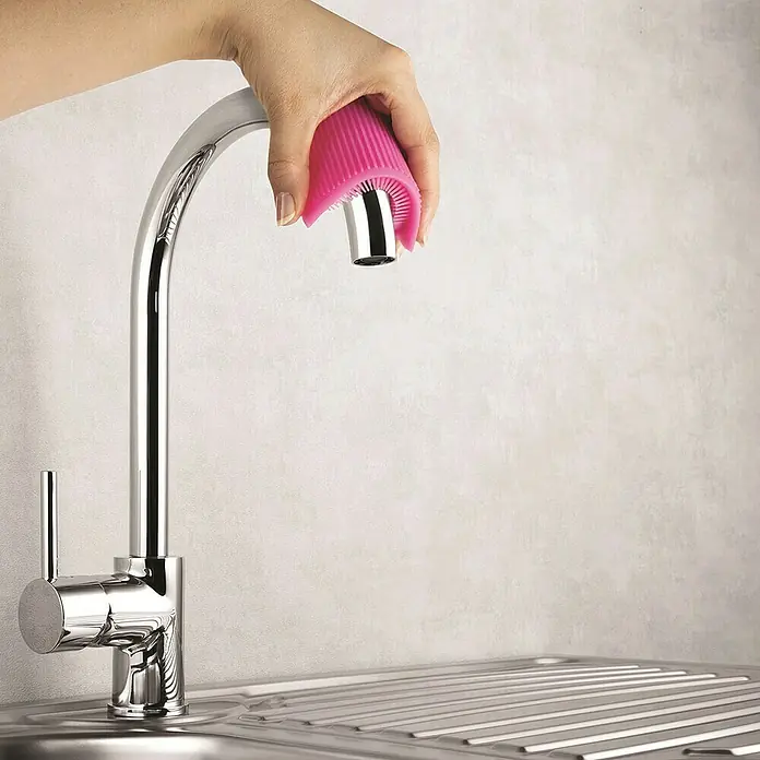Estropajo con rasqueta - 1,2 x 8,2 x 12 cm, Silicona - Primer plano de una mano que sujeta un accesorio de limpieza de silicona rosa al grifo cromado para facilitar la limpieza.